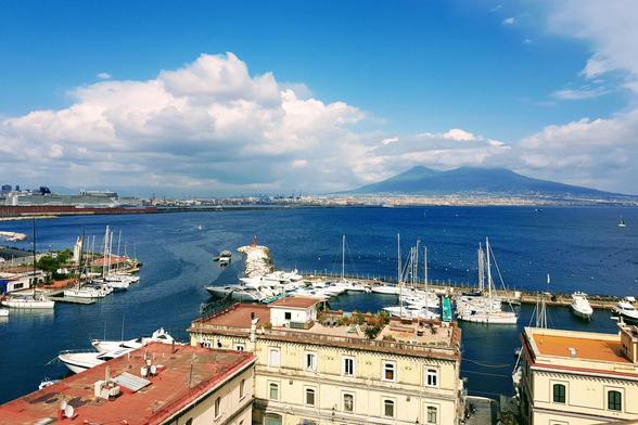 An aerial view of the Bay of Naples under a bright blue sky with fluffy white clouds. In the foreground, red-tiled rooftops of buildings line the waterfront, with a marina full of white yachts and smaller boats. The vast blue sea stretches towards the background, where Mount Vesuvius dominates the skyline. A large cruise ship is docked in the distance on the left.
