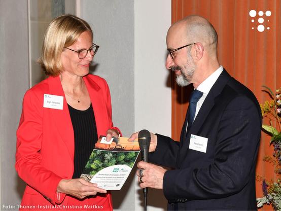 Zwei Personen stehen einander gegenüber in einem Innenraum mit heller Wand und orangefarbenem Vorhang im Hintergrund. Links im Bild steht eine Frau mit blondem Bob-Haarschnitt, Brille und rotem Blazer, die lächelt und eine Broschüre in der Hand hält. Auf ihrem Namensschild steht „Birgit Kleinschmit, Germany“. Rechts steht ein Mann mit Glatze und Brille im dunklen Anzug, der ein Mikrofon hält und freundlich auf die Frau schaut. Sein Namensschild zeigt „Marco Ferretti, Switzerland“. Die Broschüre trägt den Titel „On the Pulse of European Forests“. Im Hintergrund steht ein Blumenstrauß. Das Bild dokumentiert offenbar eine feierliche Übergabe oder Präsentation im Rahmen einer Veranstaltung.

(Foto: Thünen-Institut / Christina Waitkus)