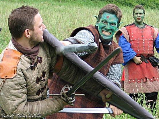 Farbbild von zwei Männern in mittelalterlichen Kostümen, die in einem inszenierten Schwertkampf gegeneinander antreten; einer von ihnen ist grün geschminkt und stellt offenbar einen Ork oder ähnliches Fabelwesen dar. Im Hintergrund beobachtet eine weitere ebenfalls grün geschminkte Person die Szene.