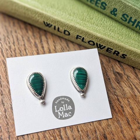 These striking green studs feature gorgeous teardrop shaped Malachite cabochons set in sterling silver, and form part of my Spring 2025 ‘Spring Greens’ special collection. The beautiful fresh green colours and patterns of the malachite are completely natural. The silver settings have a serrated edge detail and a small silver ball accent for a pretty finishing touch.

The earrings are displayed on a plain white card bearing the LollaMac logo, on a wooden surface next to some old green hardback books.