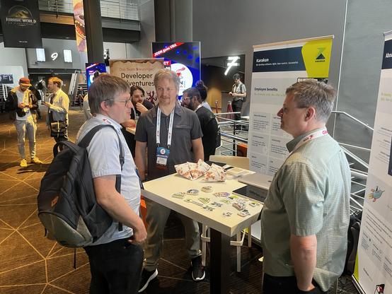 Karakun-Mitarbeiter Markus Schlichting und Marcel Rüedi im Gespräch mit einem Besucher an unserem Messestand bei der JCON 2025. Auf dem Tisch: Sticker, Flyer und Karakun-Tüten mit Basler Läckerli als Give-aways. Im Hintergrund: weitere Messestände und Konferenzteilnehmer.