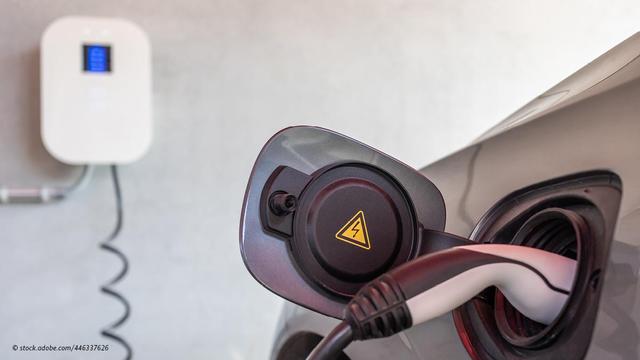 Ladeanschluss eines Elektrofahrzeuges mit eingestecktem Ladestecker. An der Wand im Hintergrund hängt eine Wallbox.