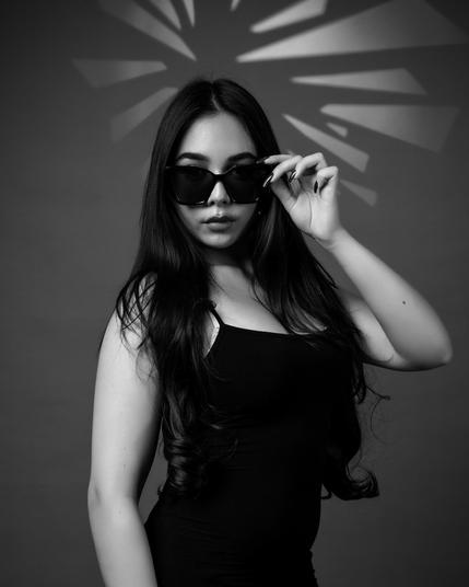 This black and white image depicts a person wearing sunglasses and a sleeveless top. They have long hair and are posing with one hand holding the glasses. The background has a decorative pattern with light rays coming out from behind.