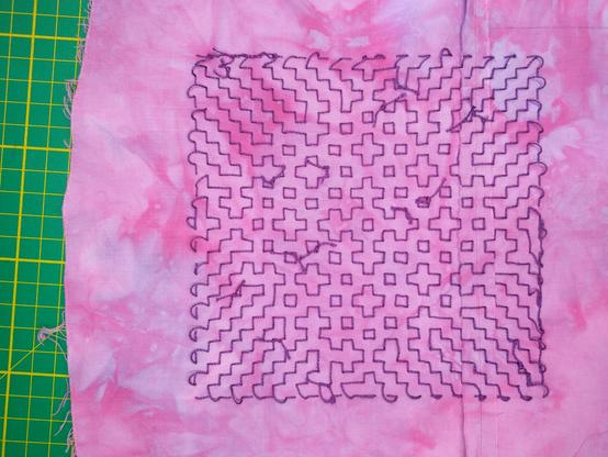 A piece of mottled dark pink/light pink fabric with a sashiko-style design embroidered on it with purple thread. It’s clearly the back of the piece, because several knots are visible where new threads have been joined in. In the middle is the order 3 Fibonacci snowflake, surrounded by small squares and Greek crosses (these, incidentally, are the order 1 and 2 snowflakes, respectively). Around the edges are zigzag lines. Like the front of the design (see previous post), the design has C4 symmetry.