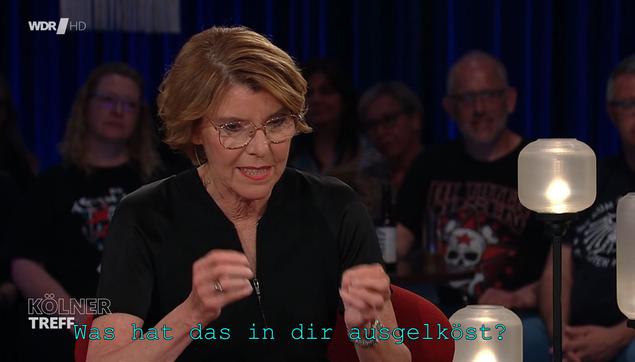 Description: The image shows a woman sitting at a table with a candle in front of her, surrounded by a group of people sitting in chairs. On the table there are jars and a lamp with a stand, and in the background there is a curtain. The woman is wearing glasses and there is text on the image.

Disclaimer: This image description was automatically generated and may not be entirely accurate.