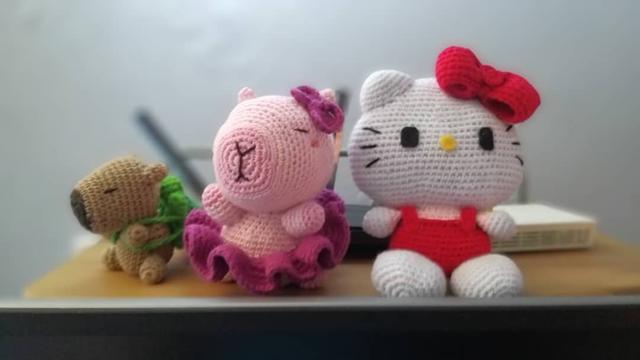 Three handmade crochet dolls sit on a wooden surface, with a blurry background of a light-colored wall and what appears to be a white electronic device. On the left is a small, light brown capybara with small ears and a green leaf-like accessory on its back. In the middle is a pink capybara wearing a ruffled dark pink skirt and a dark pink bow on its head, with its snout facing slightly to the left. On the right is a white Hello Kitty doll with black whiskers, a yellow nose, black eyes, and a prominent red bow on its head. It is wearing a red overall-style outfit. All three dolls show visible crochet stitches.