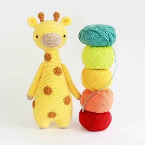 A picture of a crocheted amigurumi giraffe standing upright. It's standing next to a stack of yarn. The yarn colors from bottom to top are red, orange, yellow, green and blue.