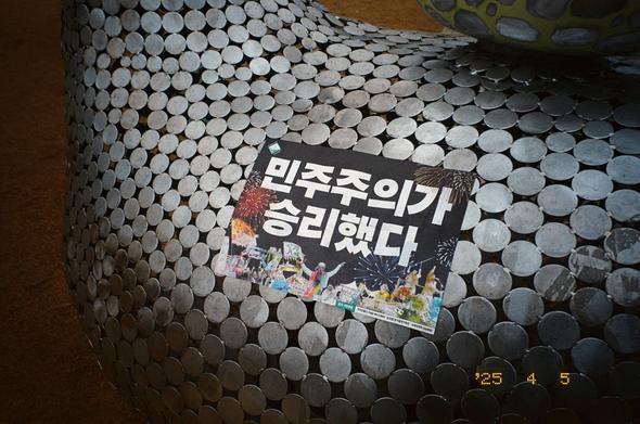 은색 동전을 이어붙인 듯한 곡선형 구조물 위에 “민주주의가 승리했다”고 쓰인 손피켓이 붙어있다. 2025-04-05