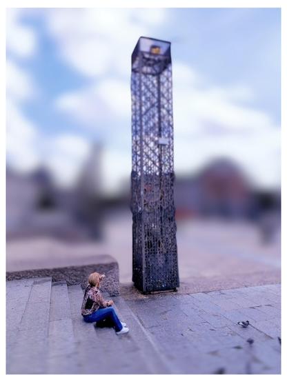 Das Bild zeigt eine Miniaturlandschaft, in der eine kleine Figur sitzt und auf eine Miniatur-Eiffeltürme-Architektur blickt. Die Figur, die auf einer Steinplattform sitzt, trägt eine Bluse mit Muster, Jeans und weiße Schuhe. Die Eiffeltürme-Architektur ist hoch und dünn, mit einem komplexen Metallgitterdesign, das die Struktur des Eiffelturms nachahmt. Der Hintergrund ist leicht verschwommen, mit einem blauen Himmel und einigen Wolken, sowie unscharfen Gebäuden im Hintergrund. Die Szene ist in einem Miniaturstil gestaltet, was die Figur und die Struktur in Bezug auf die Umgebung verkleinert.