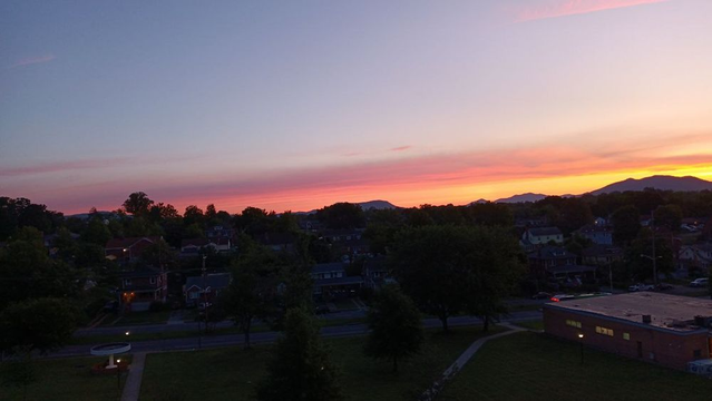 A pink and yellow and blue sunrise silhouetted by the Blue Ridge Mountains with a tree-speckled, still fairly dark urban landscape around it of houses, sidewalks, a 4 way street, and a post office prominently featured.