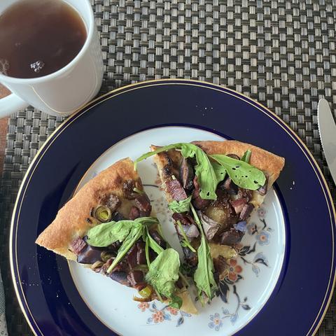 An image of two slices of pizza topped with greens and various ingredients, placed on a decorative plate. A white cup of tea is visible next to the plate