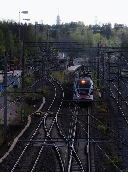 A train leaves oulunkylä station along a network of railway tracks, surrounded by trees and a television tower in the background. People are seen on the platform, and overhead electrical lines crisscross the scene.