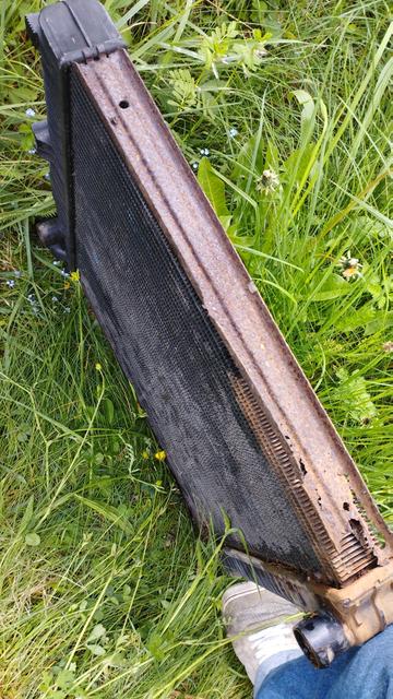 Le vieux radiateur moteur avec sa tranche bien corrodée et manquante à certains endroits. Il est posé dans l'herbe, retenu par ma jambe