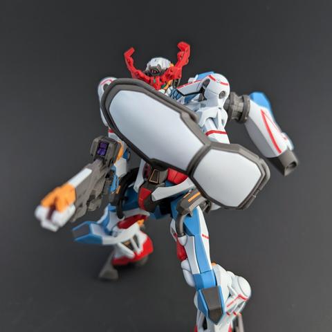 The HG GQuuuuuuX crouched down, holding a shield in front of its body and aiming its rifle at the viewer. Its left eye camera is just visible.
