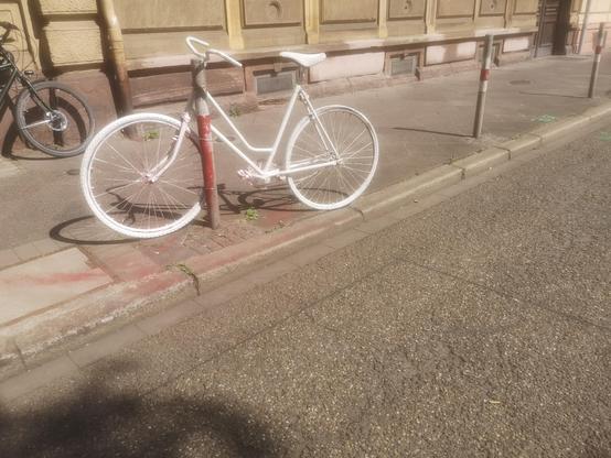 Ein weises Fahrrad lehnt an einem Poller am Straßenrand.