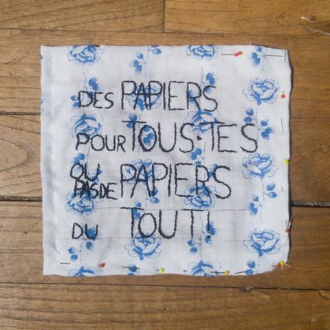 Patch brodé de " Des Papiers pour Tous•tes ou pas de Papiers du Tout !" au fil noir sur un tissu blanc avec des roses bleues.
Le patch est posé sur un plancher au bois.