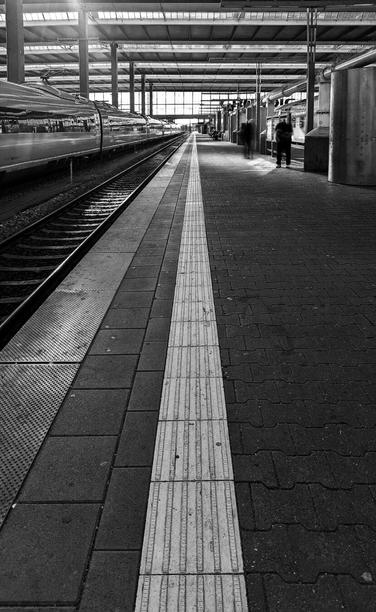 The black and white photograph captures a train platform at Munich Hauptbahnhof. The perspective is from ground level, looking down the platform. A train is visible on the left, with its tracks running parallel to the platform edge. A thick white line marks the edge of the platform. Two blurred figures are visible in the distance, suggesting movement. The platform itself is constructed from dark, rectangular paving stones. The station's roof structure is visible overhead, with sunlight filtering through.