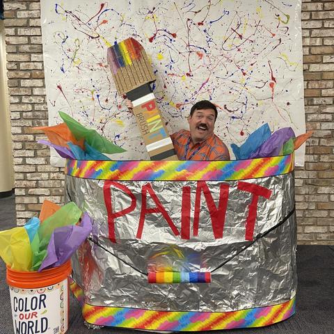A person with a mustache is playfully positioned in a large, colorful paint can, holding a giant paintbrush. Surrounding them are crumpled tissue papers in vibrant colors. Behind is a wall adorned with splattered paint for a creative backdrop