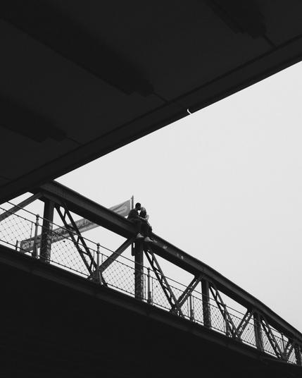This image is in black and white and depicts two people sitting on the structure of a bridge. They appear to be sitting close together on a beam above a walkway with a lattice fence. The composition includes the geometric shapes of the bridge and an overpass, creating a striking contrast against the sky.