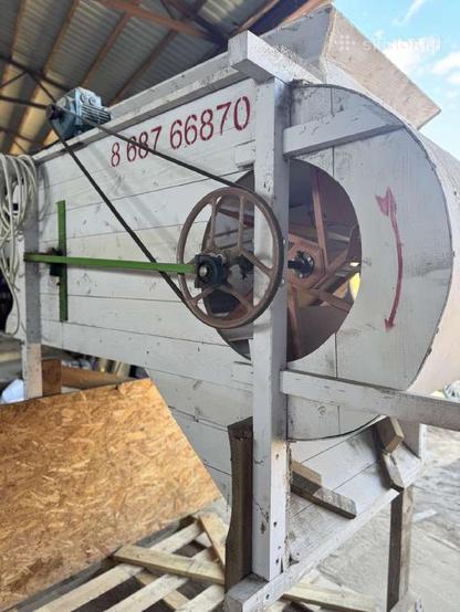 A winnowing machine, made of mostly wood with a metal fan, drive wheel and a rod linking it to the grain shaker. A belt goes to an electric motor on top. It's painted white with a red arrow indicating the direction it rotates in. A phone number is written on it for some reason.