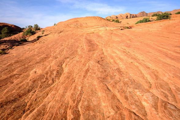 A sloping expanse of textured red sandstone with distinctive ridges and grooves, with sparse patches of green vegetation and hills in the distance under a clear blue sky.