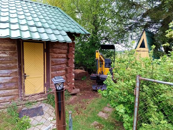 A small, yellow mini-excavator lurks in the bushes between a log sauna and an outhouse. It's driven in between two large trees in the back. A fence gate is open towards the front, just wide enough for the digger but with no space to turn. A cheap solar lamp is stuck on a fence post. A large slab of rock rests in front of the sauna door.