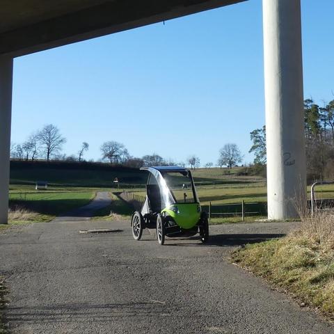 Ein Pedilio - ein Velomobil mit Solardach - steht unter einer großen Autobrücke