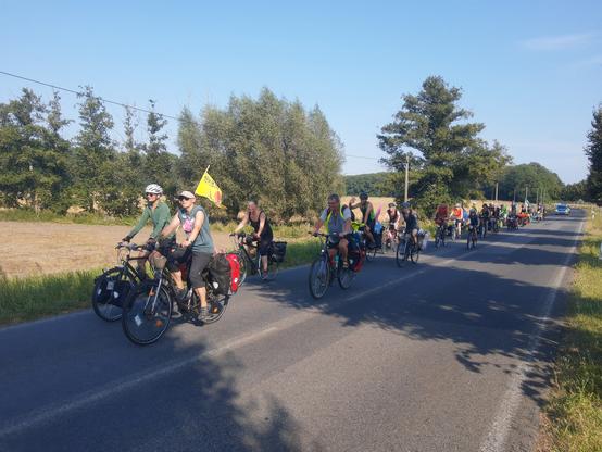 Foto von sommerlicher Landstraße mit mehreren Dutzend Radfahrenden mit Fahnen und Fahrradtaschen, ganz hinten ein Polizeiauto