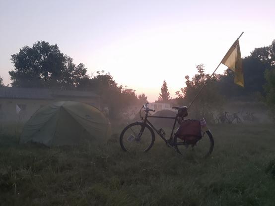 Foto eines Fahrrads mit Fahne auf einer nebligen Wiese im Sonnenaufgang, dahinter Zelte und weitere Fahrräder