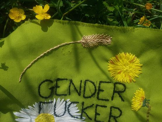Zoom sur l'épi de blé brodé sur ce patch en tissu vert. On y vois 2 pissenlits brodés dans des angles différents, le haut d'une grosse pâquerette et les mots "GENDER FUCKER"