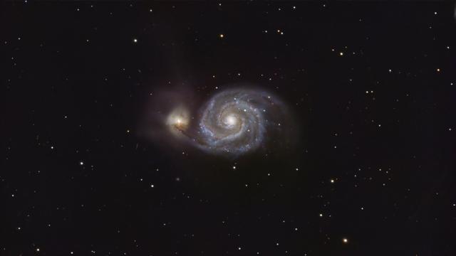 A field of stars, with a large spiral galaxy in the center. There is another lenticular galaxy to the left of it that almost looks as if it's getting sucked down into a whirlpool