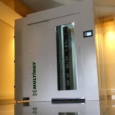 A imagem mostra um grande aparelho de laboratório branco com a marca "MULTIWAY" em destaque na lateral. O aparelho possui uma porta com uma janela de vidro que permite visualizar o interior, onde é possível ver prateleiras e equipamentos. Na parte superior da porta, há um pequeno display digital, provavelmente para controle e monitoramento. A porta possui um puxador horizontal. O aparelho está posicionado em um ambiente interno, com uma parede de cor clara ao fundo e um piso de cerâmica brilhante. A iluminação é suave, com luzes amareladas que refletem no piso, criando um ambiente limpo e organizado