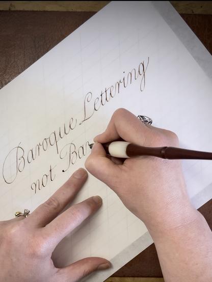 A hand is writing the phrase "Baroque Lettering not Bar…" in elaborate English Roundhand script on a piece of paper. The background features grid lines, and the hand holds a calligraphy pen. A leather surface is visible beneath the paper.