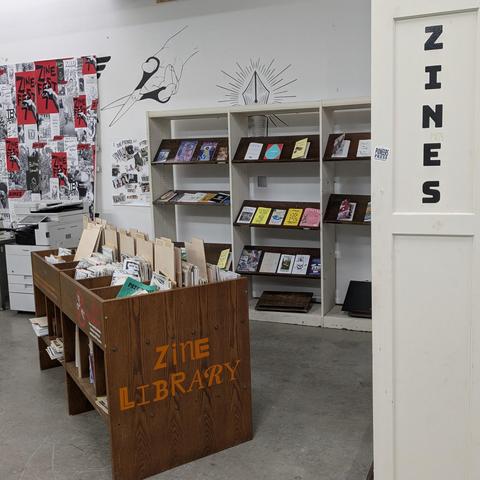 Facing a corner of the library shelves filled with old and new pamphlets labeled with "zines", "zine library", and a collage of posters for "zine fest" on the wall.