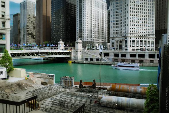 A construction site in downtown Chicago in 2005, with steel rebar and large pipes in the foreground, overlooking the turquoise waters of the Chicago River. In the background, a white riverboat cruises past the Wrigley Building (partially visible on the left), and a bridge lined with American flags spans the river. Tall skyscrapers frame the scene, and a crane hook with a 'Sox' logo hangs above the site.