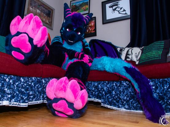 Low angle fursuit photo of Rezz sitting on a sofa showing off their footpaws