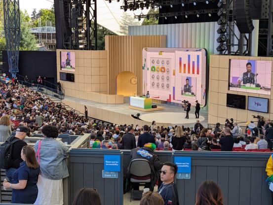 The main Google I/O stage at Shoreline from the audience. The crowd has filled in the lower level as a DJ holds the stage playing some chill tunes with Googley visualizations on screen.