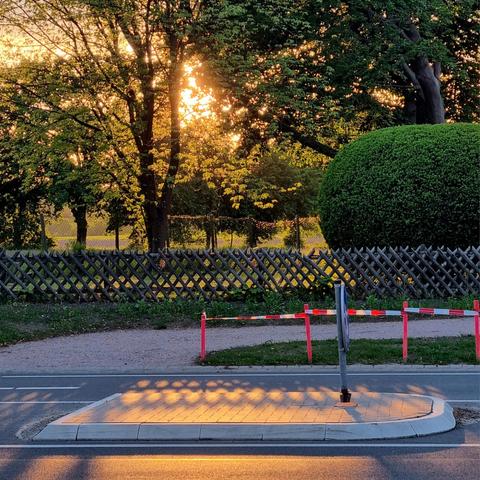 The low sun is largely hidden by trees. Rays of sunlight fall through a wooden fence with slats positioned diagonally to each other, creating wave-shaped shadows on a sunlit traffic island. At the edge of the horizontal road, a red and white striped demarcation tape provides a colorful accent to the right part of the picture.