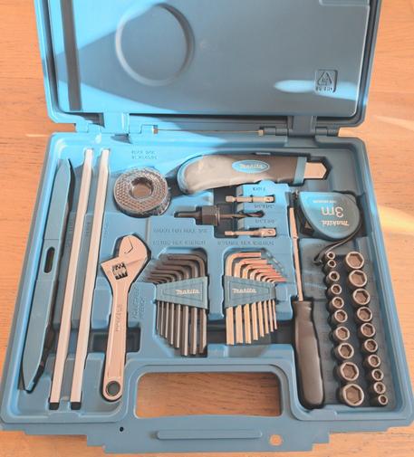 Photo lookin into the bottom compartment of a Makita box kit, containing Allen key sets, a wrench, a box cutter, a tape measure, a screwdriver with a bit holder, etc.