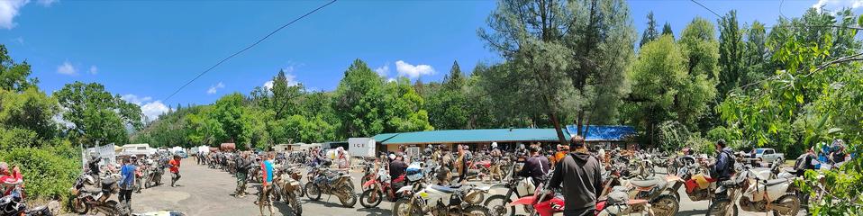 A panoramic picture of a general store / deli / fuel station at a clearing in the forest. Lots of people are parked for lunch and others waiting to buy fuel from the rusty fuel tanks on stands in the distance.