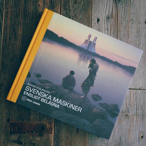 Boken ”Svenska Maskiner, Ensligt Belägna” av Simon Stålenhag. På bokens omslag syns två ungdomar vid ett vatten i solnedgången. Vid horisonten syns en stor futuristiskt byggnad. Boken ligger på en bord av trä.