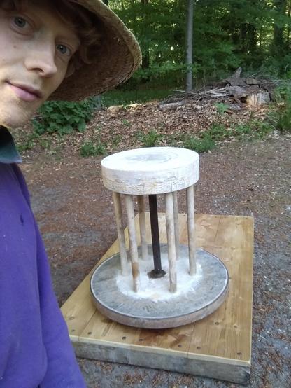A selfie with a happy looking straw hat dude in the woods. Behind him there's a true wonder of medieval technology: a potter's wheel that consists of a platform with a kickwheel on top.