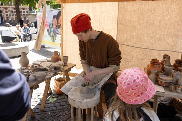 Same dude but on a market square, using the potter's wheel in medieval attire while some people are watching