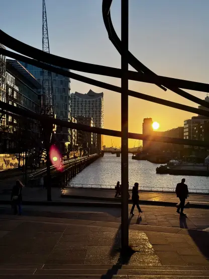 A setting sun peaking out behind some buildings in the background, partially reflecting in the fleet and the modern buildings surrounding the body of water. In the foreground, a bent rod structure, appearing black against the sunset