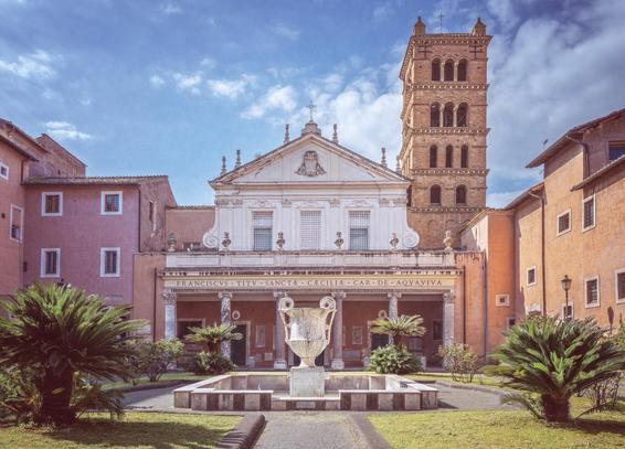 The facade and a bell tower of St Cecilia Basilica of Rome Italy in the background. In the foreground is a symmetrical courtyard with lush greenery, a fountain with a water urn in the center.