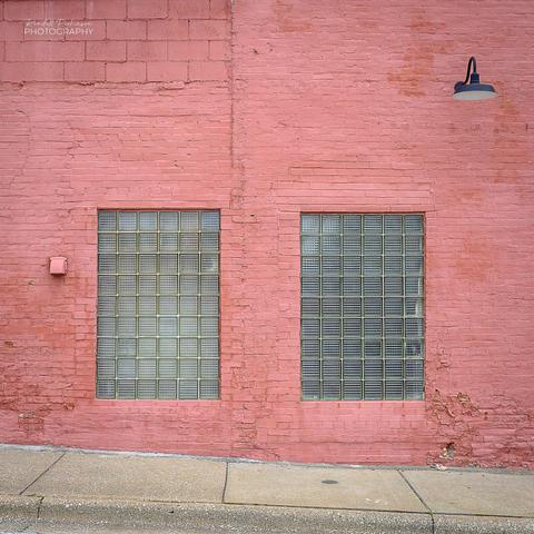 Two large glass block windows in the side of a brick building painted red.  There is an overhead light and a vent on the side of the building.