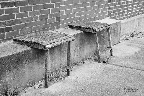 Two makeshift chairs made of seat bottoms attached to a stepped concrete foundation and legs to support the front edge.  Each is designed to fold up against the brick building so as not to obstruct the sidewalk.