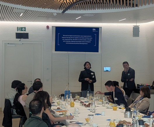 In einem Meeting-Raum des Europäischen Parlaments: Marlene Straub und Erik Tuchtfeld stehen vorne und stellen D64 vor. Marlene Straub spricht gerade. An einer langen Tafel vor ihnen sitzen Menschen beim Frühstück. An der Wand hinter den Vortragenden ist ein Fernseher mit einer Präsentation zu sehen.