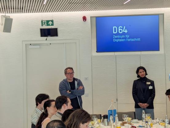 In einem Meeting-Raum des Europäischen Parlaments: Tiemo Wölken und Marlene Straub stehen vorne. Tiem Wölken spricht gerade. An einer langen Tafel vor ihnen sitzen Menschen beim Frühstück. An der Wand hinter den Vortragenden ist ein Fernseher mit dem Logo von D64 zu sehen.