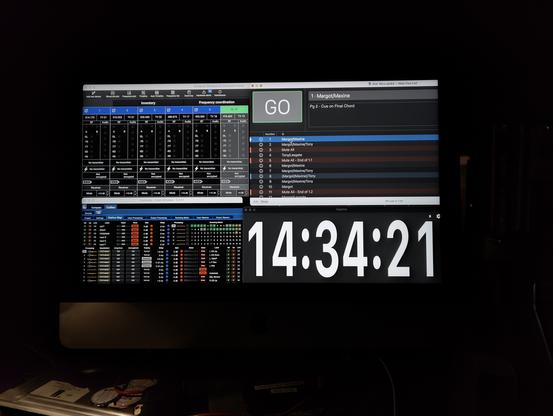 A photo of an iMac monitor displaying 4 applications roughly taking up a quarter of the screen. The top-left is Shure Wireless Workbench, to the right is Qlab. Bottom left is Meyer Compass and finally a giant digital clock called Bigtime.